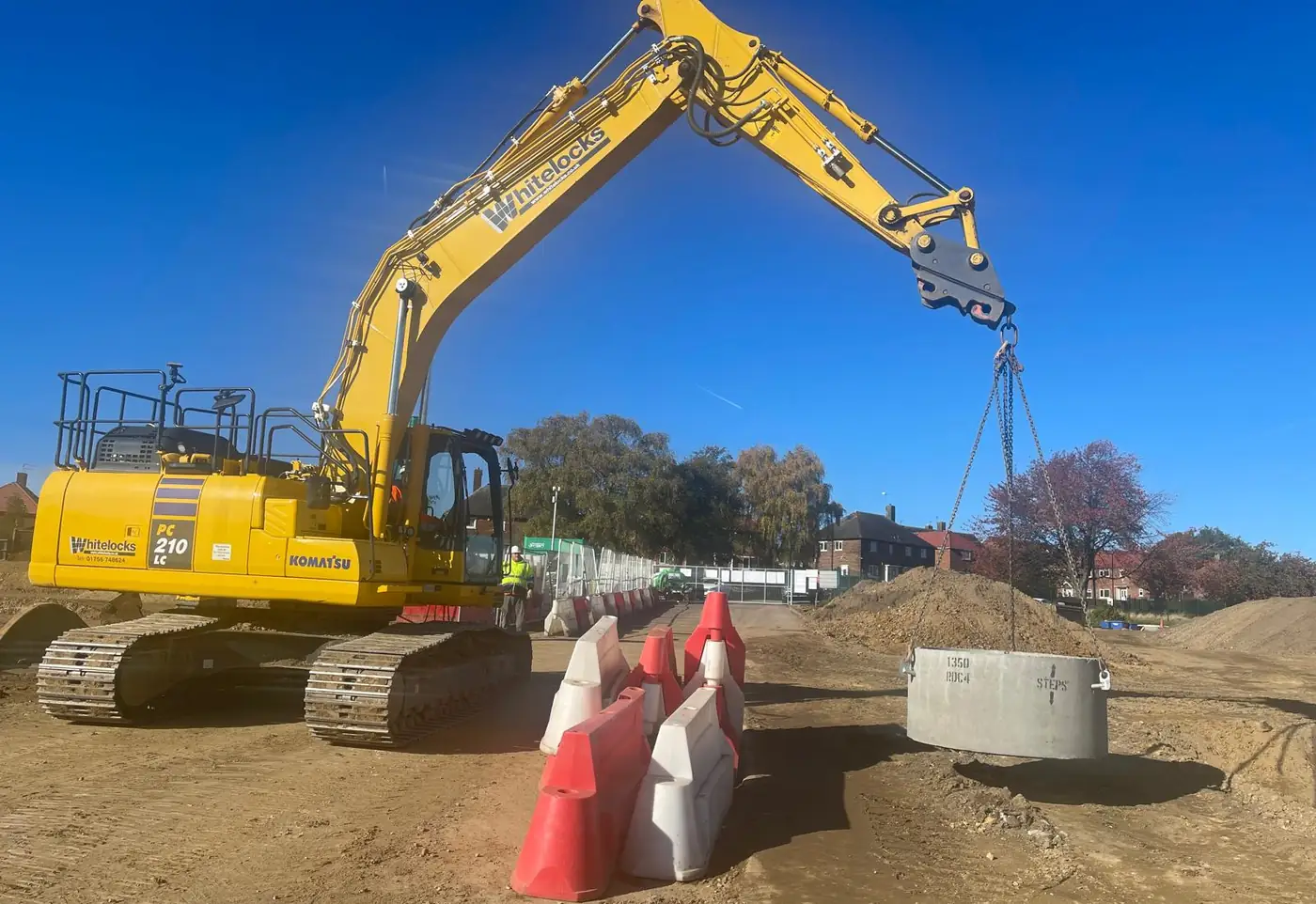 A Komatsu PC210LC 360-degree excavator lifting a concrete chamber ring on chains, with an operative in hi-vis alongside, on a live groundworks site